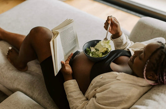 Pregnant person resting on a sofa, reading a book and eating a bowl of salad.